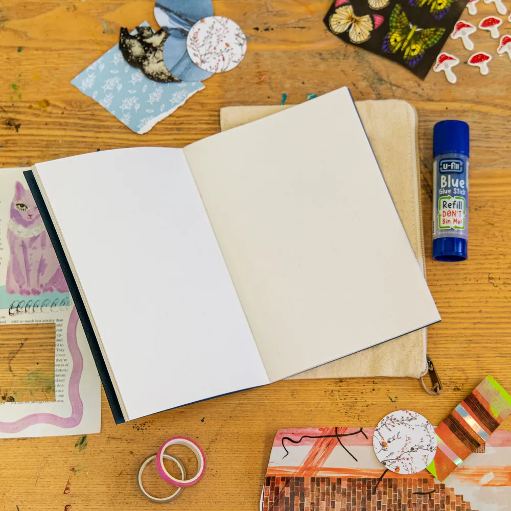 A blank art journal open on a canvas pouch, surrounded by collage scraps, stickers and washi tape.