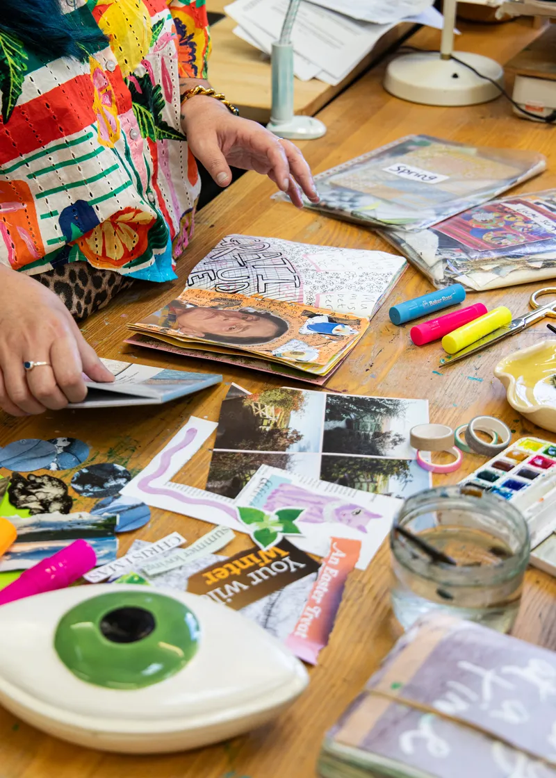 A well-used art journal open on a studio table, its pages layered with collage, drawing and handwriting.