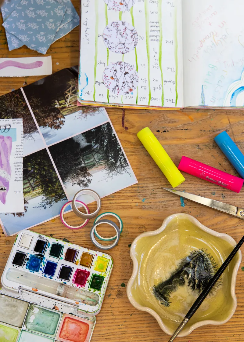 Watercolours, glue sticks, washi tape and scissors laid out beside an open journal.