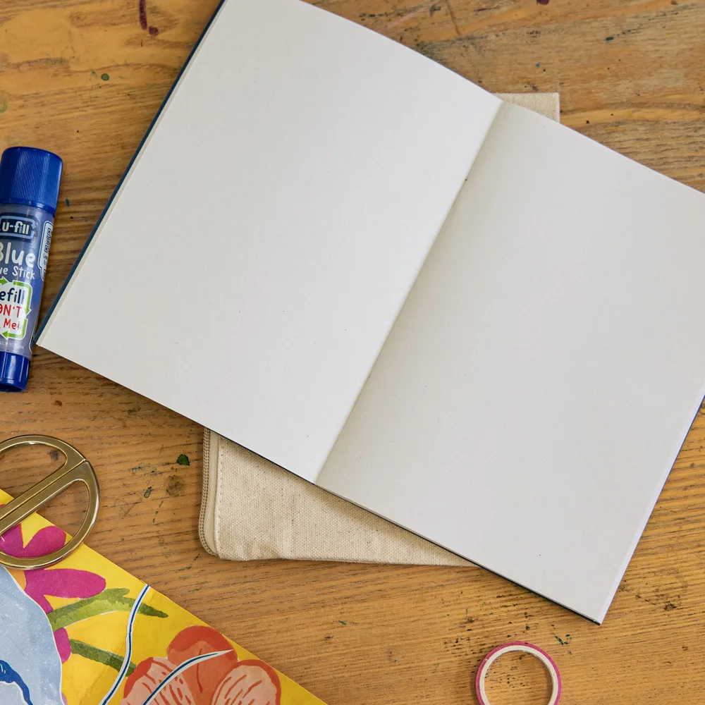 An open, blank art journal on a canvas pouch, with a glue stick, gold scissors, washi tape and colourful collage paper on a paint-splattered studio table.