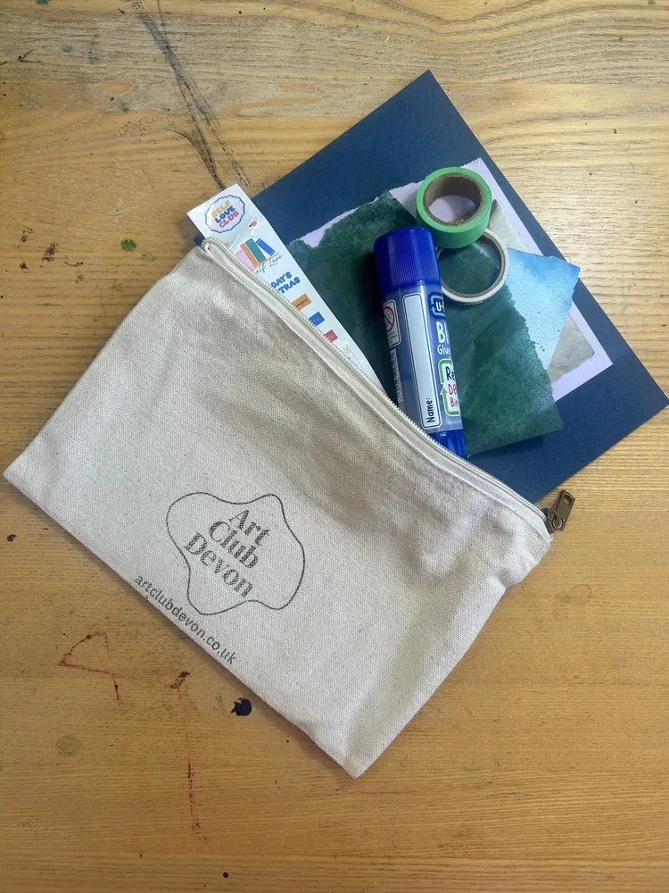 The Art Club collage kit pouch, with the journal, glue stick, washi tape, and stickers peeking out of it, on a paint-splattered studio table.